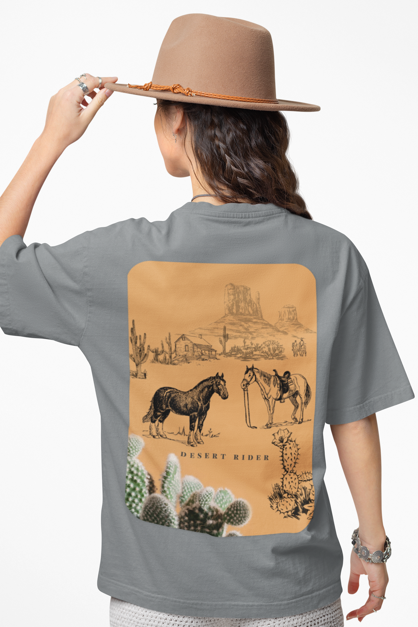 Person wearing a gray t-shirt with a desert-themed graphic on the back, holding a brown hat.