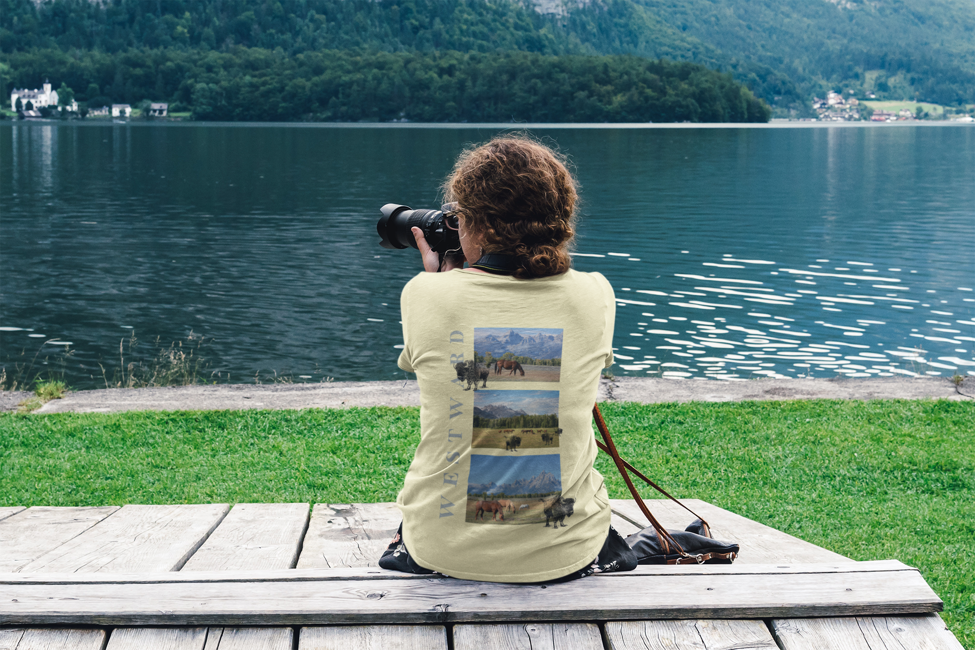 Person sitting by a lake, taking photos with a camera, wearing a t-shirt with nature-themed design.