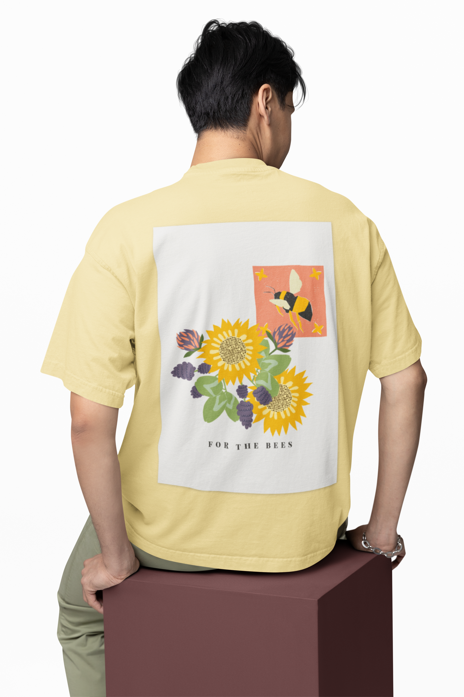 Person wearing a light yellow t-shirt with a floral design on the back, sitting on a brown box against a white background.