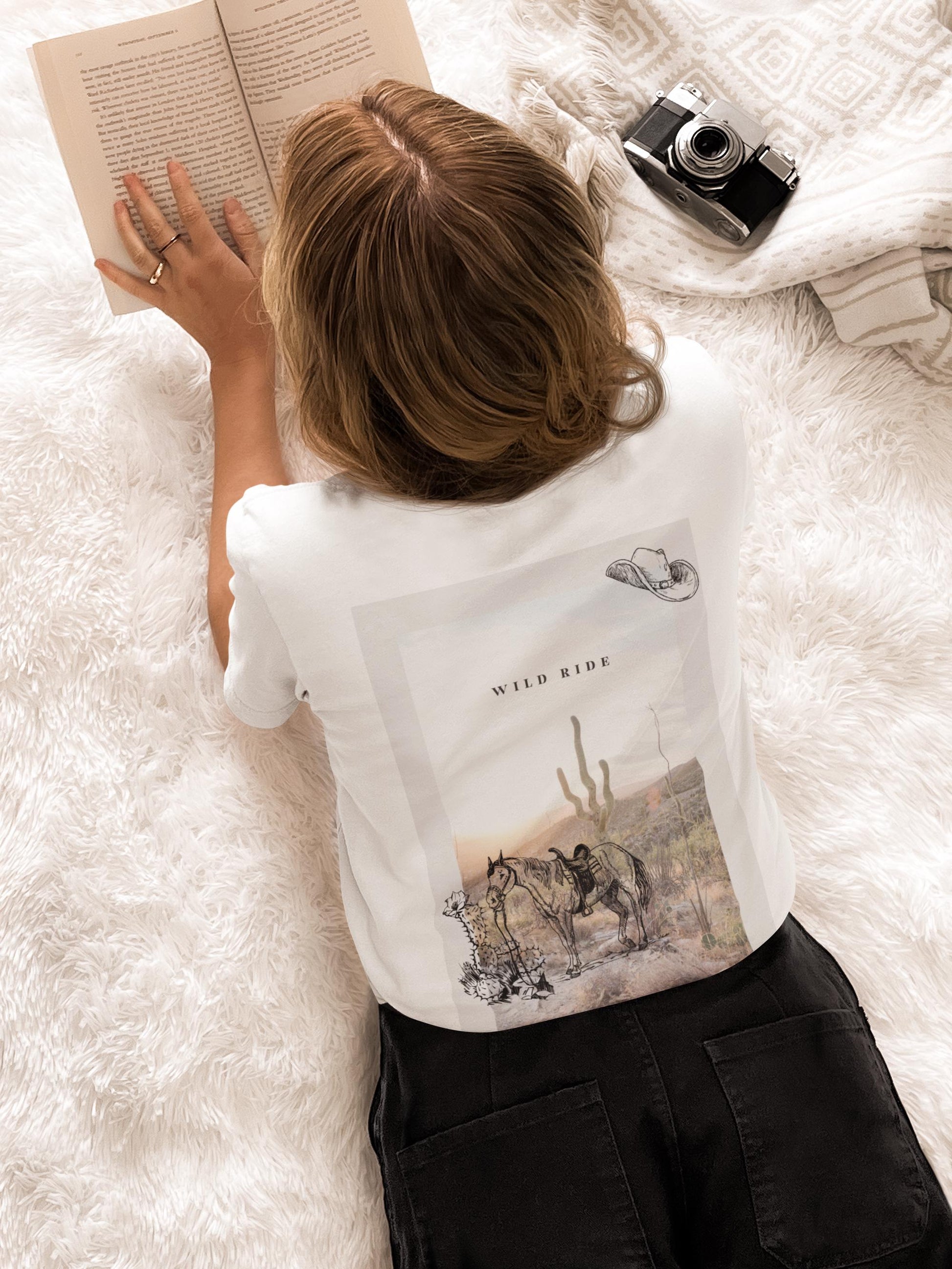 Person lying on a white surface reading a book with a camera and white shirt with graphic design.