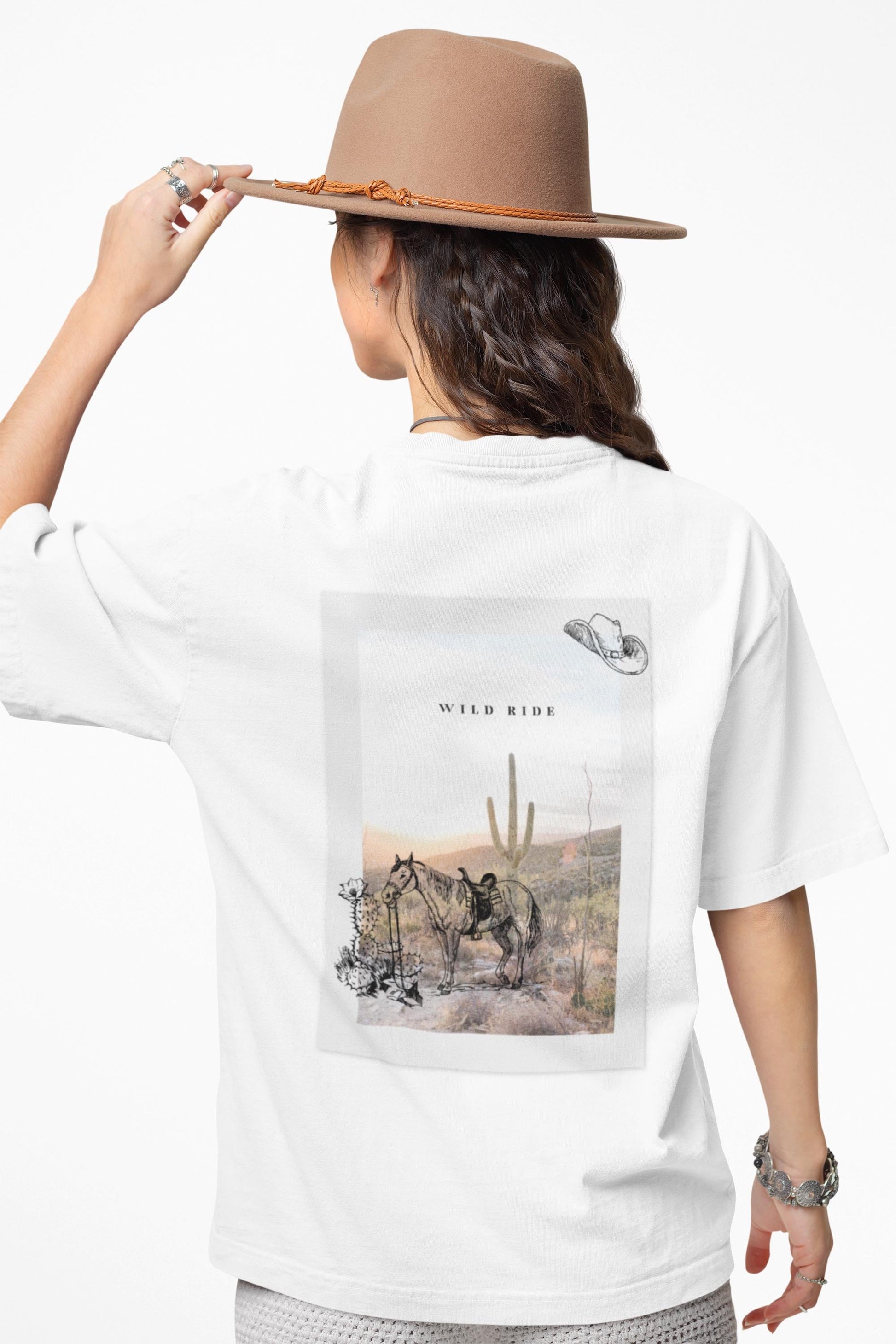 Person wearing a white t-shirt with a desert scene design, holding a beige hat.